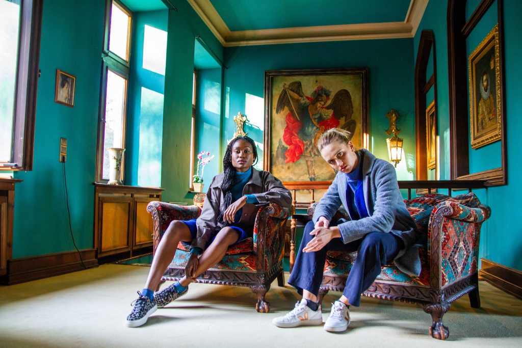 Zwei Models in modernen Fashion Outfits in einem türkisen Raum, dynamisch inszeniert von Fotografin Malin Kalwa für Fashion Content