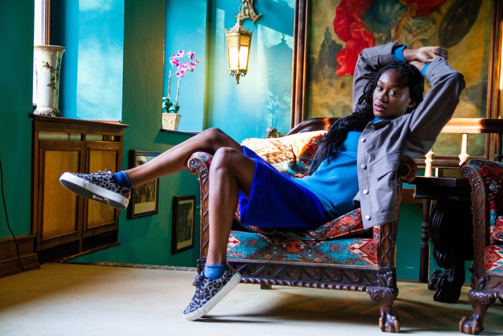 Fashion-Model in blauem Rock und grauer Jacke, lässig auf Sessel posierend, Foto von Malin Kalwa für Mode-Editorial