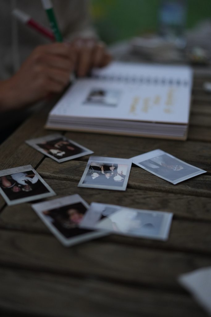 Detailaufnahme von Sofortbildern (Polaroids) auf einem Holztisch, während im Hintergrund eine Person Notizen macht – Erinnerungs- und Hochzeitsfotografie Bonn.