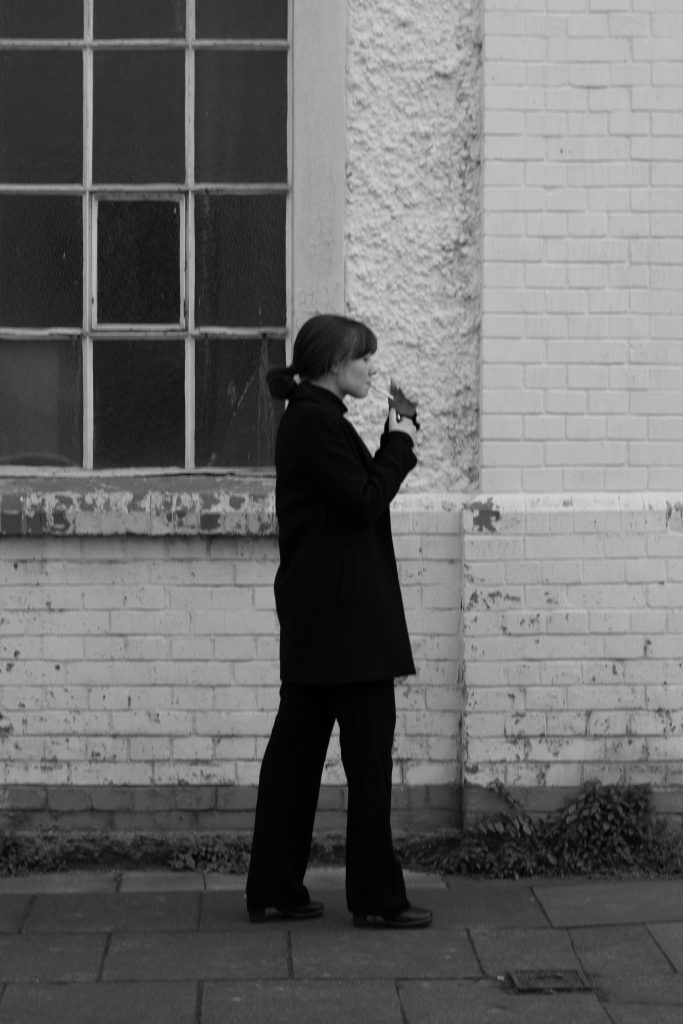 Minimalistisches Fashion-Foto einer Frau im Profil mit schwarzem Mantel vor Ziegelwand, erstellt von Malin Kalwa in Bonn