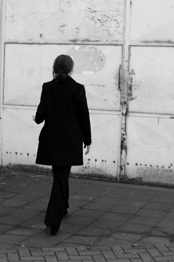 Rückenansicht einer Frau in schwarzem Mantel vor einer Industrie-Wand, stilvolle Fotografie der Bonnerin Malin Kalwa